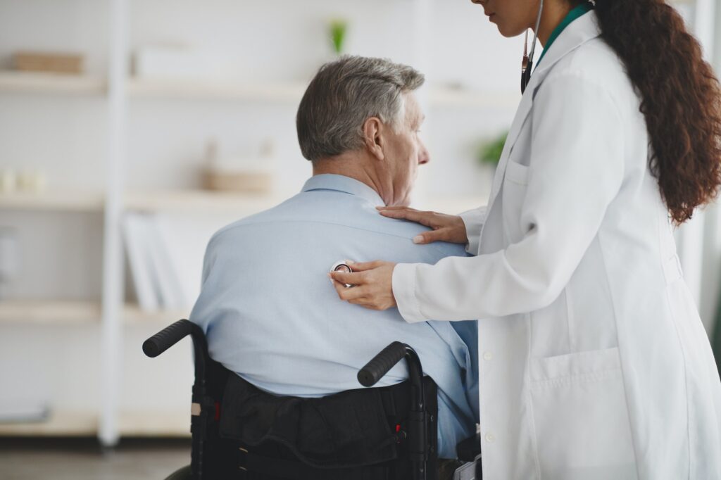 Female doctor using stethoscope on older man in wheelchair at home
